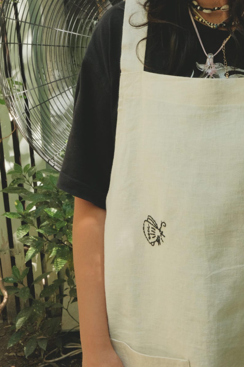 May include: An off-white apron featuring a small, black embroidered butterfly design on the front. The apron has wide shoulder straps and is worn over a black short-sleeved shirt. The fabric has a visible weave texture throughout the garment.
