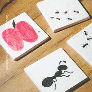 Pu&ograve; includere: Quattro sottobicchieri quadrati bianchi con illustrazioni in bianco e nero. Uno mostra una mela rossa con picciolo nero, un altro una grande formica nera e due mostrano diverse piccole formiche nere. Il testo MOOBI &egrave; stampato in basso.