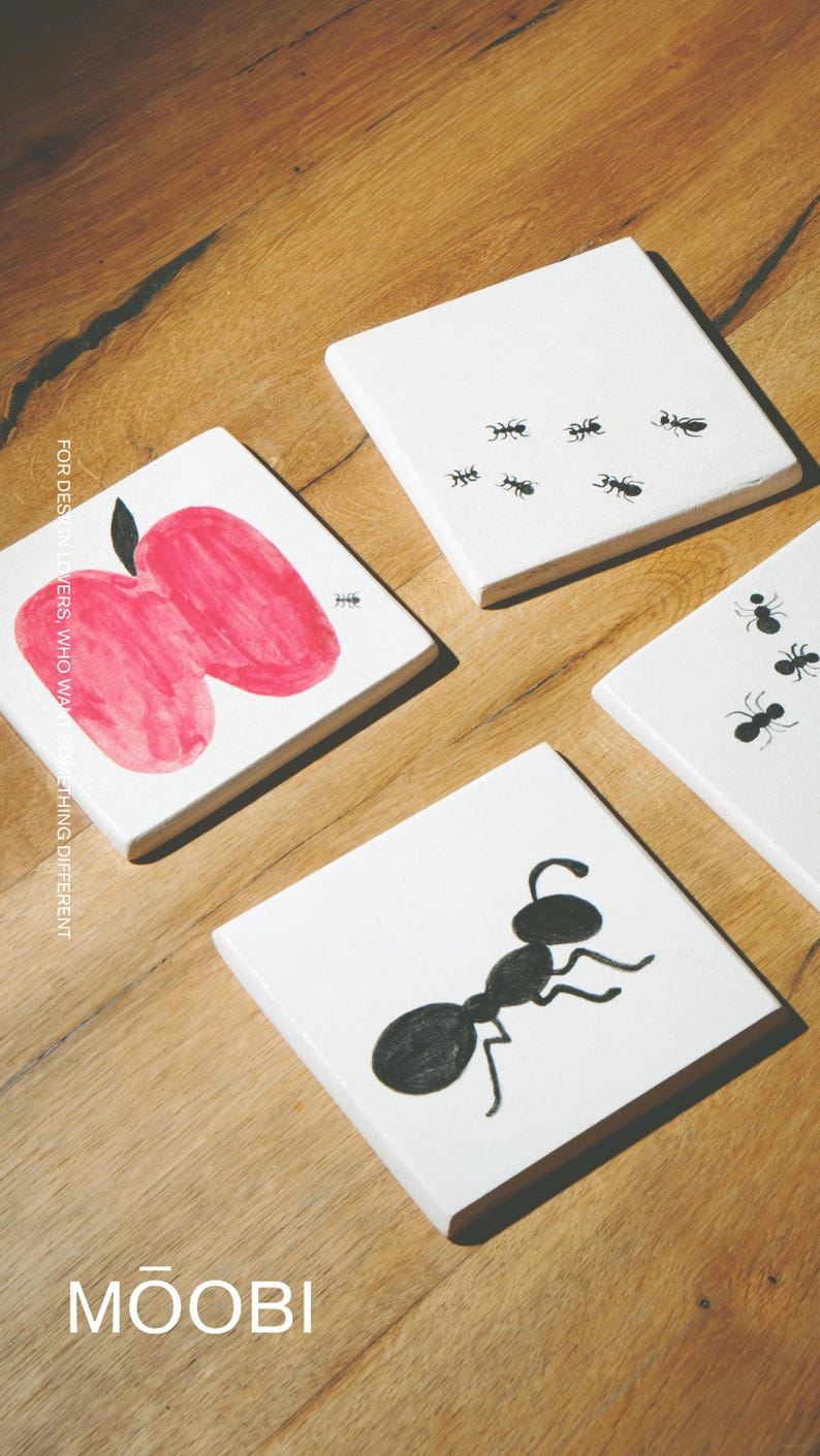 Pu&ograve; includere: Quattro sottobicchieri quadrati bianchi con illustrazioni in bianco e nero. Uno mostra una mela rossa con picciolo nero, un altro una grande formica nera e due mostrano diverse piccole formiche nere. Il testo MOOBI &egrave; stampato in basso.