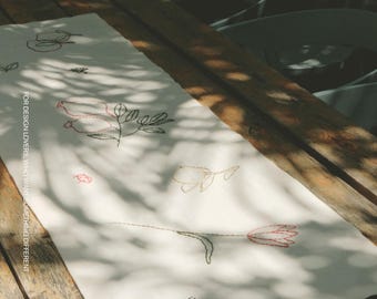 Puede incluir: Un camino de mesa rectangular de color blanco roto con bordados de líneas finas de varios elementos botánicos y pequeños insectos en hilo negro y rojo. El texto MOOBI y FOR DESIGN LOVERS, WHO WANT SOMETHING DIFFERENT está impreso en él.