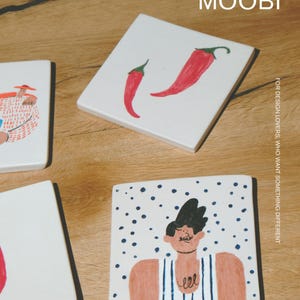 May include: Three square white ceramic tiles featuring hand-painted illustrations. One tile displays two red chili peppers, another shows a person in a striped tank top against blue dots, and the third shows a person in a hat. Text reads MŌOBI.