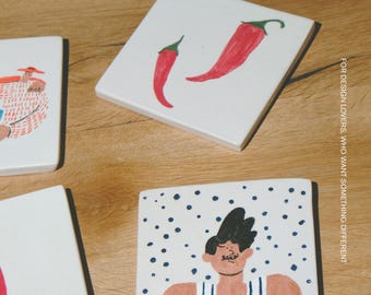 Puede incluir: Tres azulejos de cerámica blancos cuadrados con ilustraciones pintadas a mano. Un azulejo muestra dos chiles rojos, otro una persona con camiseta de tirantes a rayas sobre puntos azules, y el tercero una persona con sombrero. El texto dice MŌOBI.