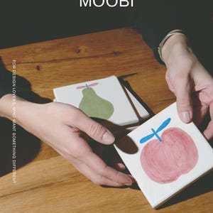 Puede incluir: Posavasos cuadrados de cer&aacute;mica blanca con ilustraciones de frutas pintadas a mano. Un posavasos muestra una pera verde con un tallo azul, mientras que otro muestra una manzana rosa con un tallo azul. El texto MŌOBI y FOR DESIGN LOVERS, WHO WANT SOMETHING DIFFERENT son visibles.
