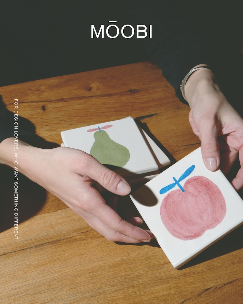 Puede incluir: Posavasos cuadrados de cer&aacute;mica blanca con ilustraciones de frutas pintadas a mano. Un posavasos muestra una pera verde con un tallo azul, mientras que otro muestra una manzana rosa con un tallo azul. El texto MŌOBI y FOR DESIGN LOVERS, WHO WANT SOMETHING DIFFERENT son visibles.