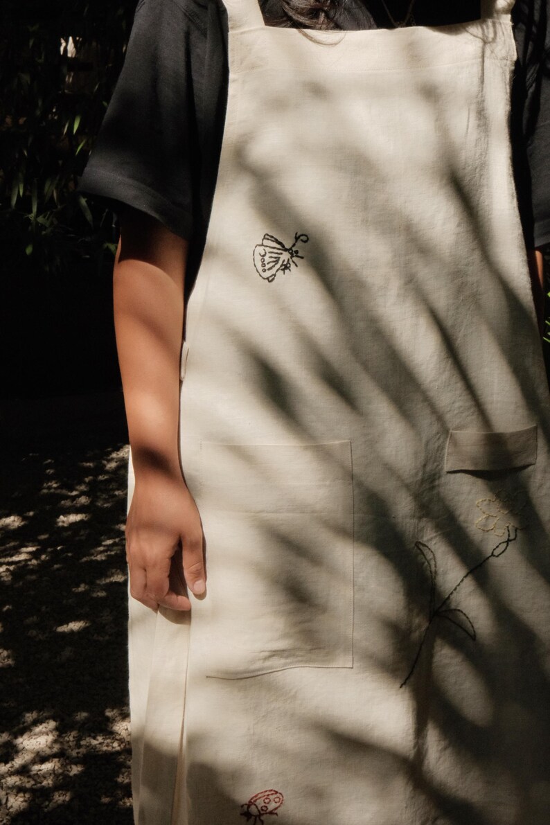 May include: An off-white apron featuring embroidered details including a black moth, a small red ladybug, and a floral stem. The apron has a square neckline with shoulder straps and a large front pocket, crafted from a plain woven fabric.