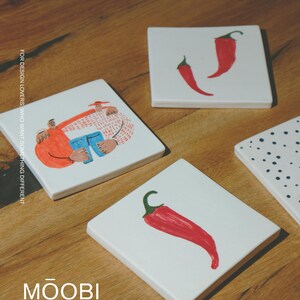 May include: Four square white ceramic coasters featuring hand-painted illustrations. One shows two figures embracing, two feature red chili peppers, and one has black polka dots. The text reads FOR DESIGN LOVERS, WHO WANT SOMETHING DIFFERENT and MŌOBI.