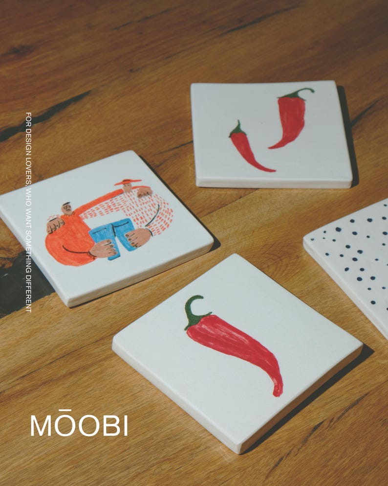 May include: Four square white ceramic coasters featuring hand-painted illustrations. One shows two figures embracing, two feature red chili peppers, and one has black polka dots. The text reads FOR DESIGN LOVERS, WHO WANT SOMETHING DIFFERENT and MŌOBI.