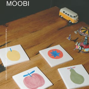 Puede incluir: Cuatro posavasos cuadrados de cer&aacute;mica blanca con ilustraciones de frutas pintadas a mano: una fruta amarilla, un c&iacute;rculo rojo con centro azul, una manzana rosa con tallo azul y una pera verde. El texto MŌOBI y FOR DESIGN LOVERS WHO WANT SOMETHING DIFFERENT son visibles.