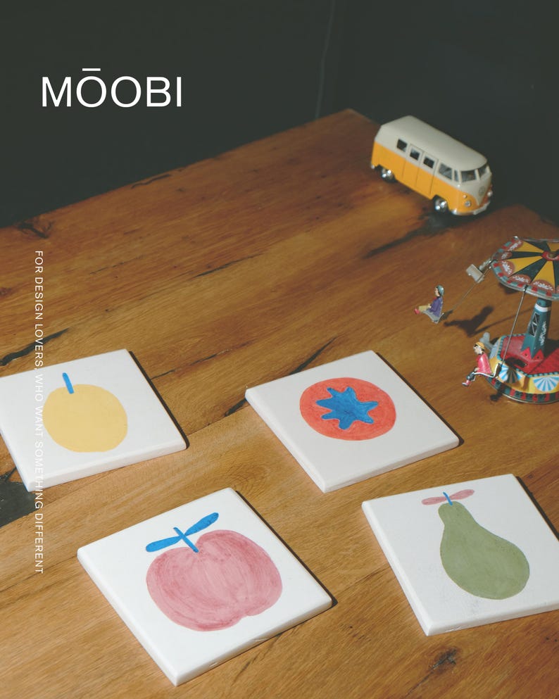 Puede incluir: Cuatro posavasos cuadrados de cer&aacute;mica blanca con ilustraciones de frutas pintadas a mano: una fruta amarilla, un c&iacute;rculo rojo con centro azul, una manzana rosa con tallo azul y una pera verde. El texto MŌOBI y FOR DESIGN LOVERS WHO WANT SOMETHING DIFFERENT son visibles.