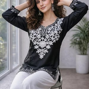 Puede incluir: Una túnica negra de manga larga con escote en V, adornada con intrincados bordados florales blancos en el pecho y los puños. La prenda incluye un borde bordado en blanco a lo largo del dobladillo y se combina con pantalones blancos lisos.