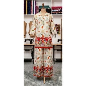 Könnte beinhalten: Ein zweiteiliges Outfit bestehend aus einer langärmeligen Tunika und einer weiten Hose. Der Stoff hat eine cremefarbene Basis mit einem sich wiederholenden geometrischen Muster in Grün und Orange, akzentuiert durch einen roten Rand mit detaillierten figürlichen Illustrationen.