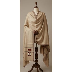 Op de afbeelding: Een beige sjaal met een donkerbruine rand en dunne horizontale strepen bij de zoom. De sjaal is voorzien van een rechthoekig label met de tekst "PASHMINA" en "100% CASHMERE" in wit en rood, gedrapeerd over een houten paspopstandaard.