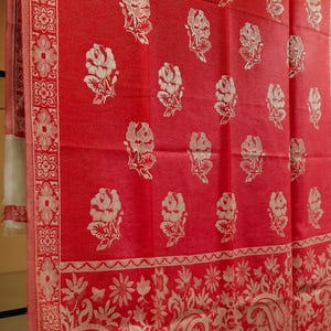 May include: A vibrant red shawl featuring a repeating pattern of cream-colored roses across the main body. The wide border displays intricate floral and paisley motifs in cream. A label at the bottom corner reads Oswal Shawls & Lohi.