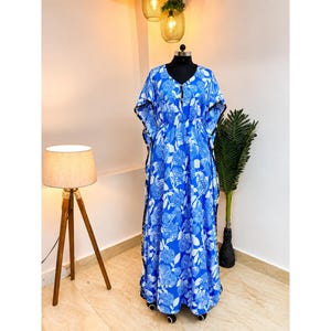 May include: A floor-length kaftan dress featuring a vibrant blue and white floral pattern. The garment has a V-neckline, loose kimono-style sleeves, and a cinched waistline. The edges of the sleeves and sides are finished with a thin black trim.
