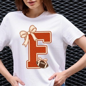 Pode incluir: Uma t-shirt branca de manga curta com uma letra F laranja texturizada grande e um contorno cor de creme. A letra é decorada com um laço cor de creme no canto superior esquerdo e uma pequena bola de futebol americano castanha com um laço a condizer.