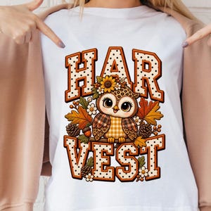 May include: A white t-shirt featuring a graphic of a cartoon owl with large eyes, wearing a sunflower on its head. The owl is surrounded by autumn leaves, pinecones, and flowers. The word HARVEST is printed in large, orange, polka-dot letters.