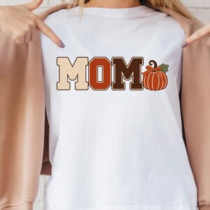 May include: A white t-shirt featuring the text MOM in a block font with a gradient of beige, orange, and brown colors. The letter O is replaced by an orange pumpkin illustration with a green leaf and a small bow tied to its stem.