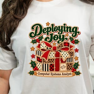 Può includere: Una t-shirt bianca con la grafica di un pacco regalo con un grande fiocco rosso, circondato da foglie di agrifoglio, bacche e stelle. Il testo recita "Deploying Joy" in alto e "Computer Systems Analyst" in basso con un simbolo di codice.