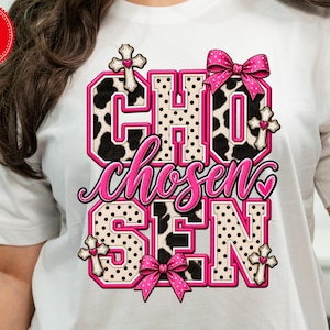 Puede incluir: Una camiseta blanca con un diseño gráfico que muestra la palabra CHOSEN en letras grandes. Las letras alternan entre estampado de vaca en blanco y negro y lunares beige, decoradas con lazos rosas, pequeñas cruces y corazones rosas.