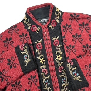 May include: A red and black knit zip-up jacket featuring a repeating black floral pattern across the body. The front placket, collar, and cuffs are trimmed in black with embroidered red, yellow, and blue floral vines. The label reads Icelandic Design.