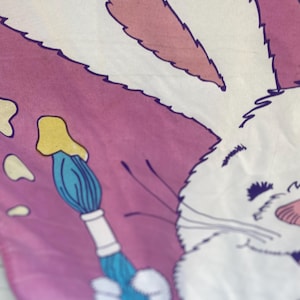 May include: A white and pink Easter-themed fabric with a cartoon bunny holding a paintbrush. The bunny has white fur, pink ears, and a pink nose. The paintbrush is blue with a white handle, and there are yellow paint splatters.