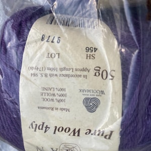 May include: A ball of deep purple Rowan Pure Wool 4ply yarn encased in clear plastic packaging. The label specifies 50g, 174 yards, 100% wool, lot 9778, and shade 456. The yarn is made in Romania and features the Woolmark logo on the white paper label.