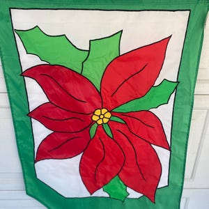 May include: A green and white Christmas flag featuring a large red poinsettia flower with green leaves. The flower has a yellow center. The flag has a green border and is designed for outdoor display.