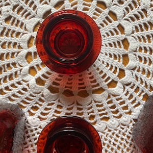 May include: Two deep red glass candle holders viewed from directly above. Each holder features a circular opening with a thick, smooth rim and a textured, faceted exterior surface. The glass is translucent, allowing light to pass through the rich crimson hue.