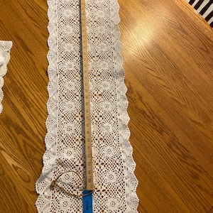 May include: A long, rectangular white crochet table runner featuring an intricate floral pattern and a scalloped edge. A wooden yardstick is placed vertically along the center, showing the runner measures approximately 36 inches in total length.