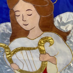 May include: A fabric angel with brown hair, closed eyes, and a gold halo. The angel wears a white robe and holds a gold harp. The background is blue with white and silver accents. A red ribbon is visible.
