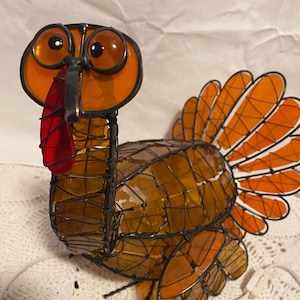 May include: A decorative turkey figurine made of orange and amber-colored glass pieces, framed by a dark metal wire. The turkey has large orange eyes, a red wattle, and a fanned tail. The figurine is designed to be a Thanksgiving decoration.