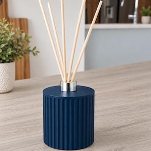 May include: A cylindrical reed diffuser with a deep navy blue, vertically ribbed exterior. The top features a metallic silver collar holding five light-coloured wooden reeds that extend upwards from the centre of the vessel.