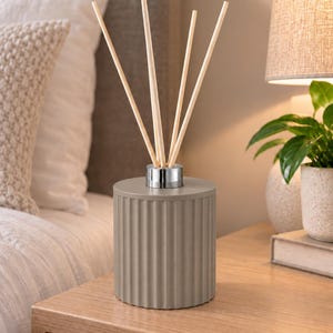 May include: A cylindrical reed diffuser with a ribbed, matte taupe exterior and a polished silver-tone metal collar at the top. Four light-coloured wooden reeds are inserted into the centre opening, extending upwards from the vessel.
