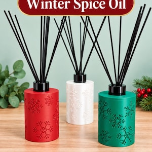 May include: Three cylindrical reed diffusers in red, white, and green, each featuring embossed snowflake patterns and black reeds. A red banner at the top displays the text Includes Winter Spice Oil with snowflake icons on both sides.