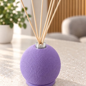 May include: A spherical purple reed diffuser with a textured surface and a silver-toned metal collar at the top. Five light-coloured wooden reeds are inserted into the centre opening of the diffuser, extending upwards from the round base.