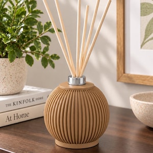 May include: A spherical reed diffuser with a ribbed tan exterior and a silver-toned metal collar at the top. Six light-coloured wooden reeds are inserted into the centre of the diffuser, extending upwards in a fan-like arrangement.