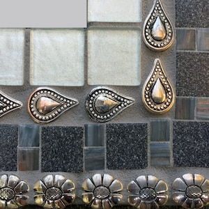 K&ouml;nnte beinhalten: Nahaufnahme eines Mosaik-Spiegelrahmens in Silber und Schwarz. Der Rahmen ist mit silbernen tropfenf&ouml;rmigen Verzierungen und silbernen bl&uuml;tenf&ouml;rmigen Verzierungen verziert.
