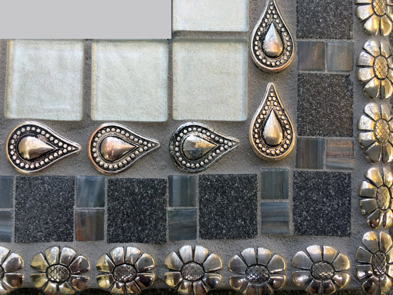 K&ouml;nnte beinhalten: Nahaufnahme eines Mosaik-Spiegelrahmens in Silber und Schwarz. Der Rahmen ist mit silbernen tropfenf&ouml;rmigen Verzierungen und silbernen bl&uuml;tenf&ouml;rmigen Verzierungen verziert.