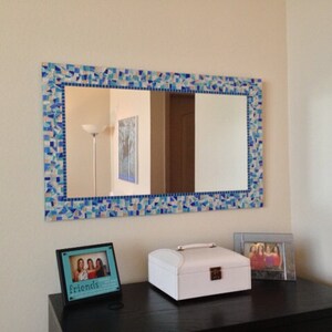 May include: A rectangular mirror with a blue and white mosaic tile frame. The mirror is hanging on a light beige wall above a black dresser. There are two picture frames on the dresser, one with a photo of three people and the text "friends" and the other with a white jewellery box.
