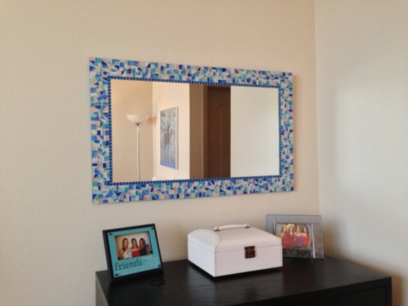 May include: A rectangular mirror with a blue and white mosaic tile frame. The mirror is hanging on a light beige wall above a black dresser. There are two picture frames on the dresser, one with a photo of three people and the text "friends" and the other with a white jewellery box.