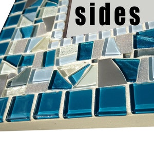 May include: Close-up of a mosaic mirror frame with blue, grey, and white tiles. The frame is made of small, irregular pieces of glass and has a rough, textured surface. The word "sides" is visible on the mirror.