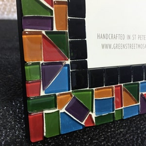 May include: A mosaic picture frame with a black border and colorful glass tiles. The frame features a mix of red, orange, green, purple, and blue tiles. The text "HANDCRAFTED IN ST PETERSBURG, FL" and a website address are visible.