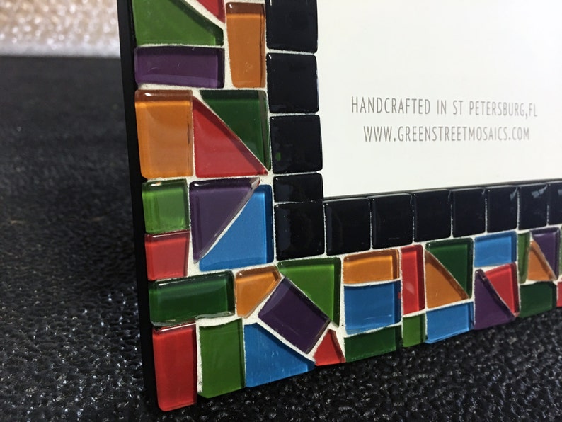 May include: A mosaic picture frame with a black border and colorful glass tiles. The frame features a mix of red, orange, green, purple, and blue tiles. The text "HANDCRAFTED IN ST PETERSBURG, FL" and a website address are visible.