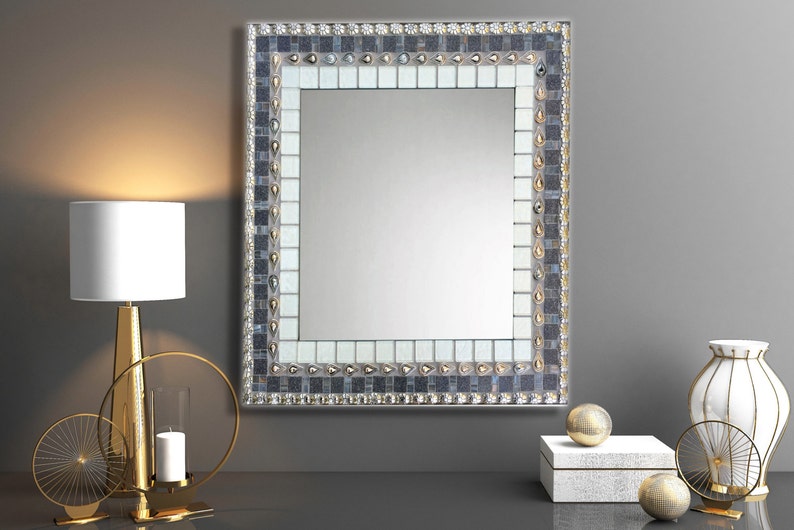 A wall mirror with a mosaic frame. The frame is made of small, square tiles in shades of gray, white, and silver.