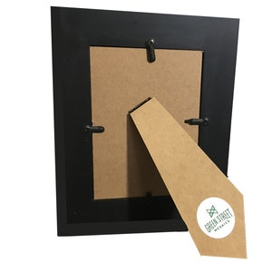 May include: A black picture frame with a brown backing and a cardboard easel. The easel has a white sticker with the text "GREEN STREET MOSAICS" in green and black. The frame is square and has a simple design.