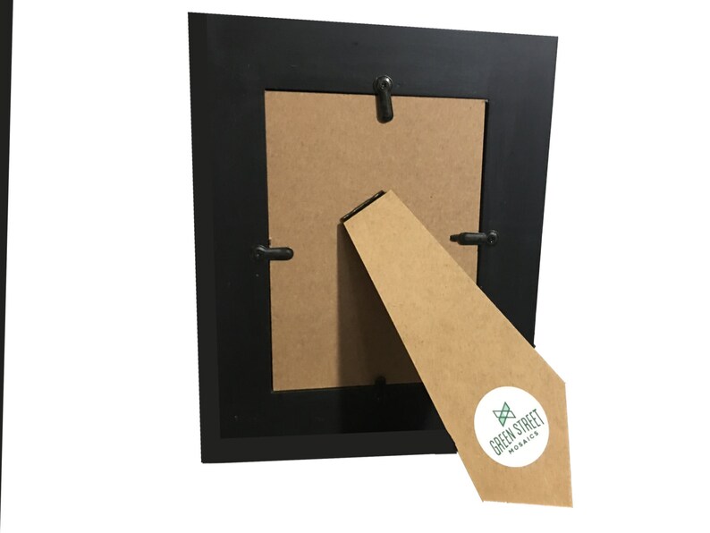May include: A black picture frame with a brown backing and a cardboard easel. The easel has a white sticker with the text "GREEN STREET MOSAICS" in green and black. The frame is square and has a simple design.