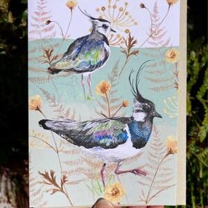May include: A greeting card featuring a watercolor illustration of two birds with long black crests and colorful plumage standing in a field of green and brown foliage with yellow flowers.