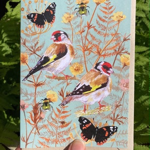 May include: A greeting card with a blue background featuring two goldfinches, two butterflies, and a bumblebee. The birds are perched on branches with green leaves and brown stems. The butterflies are flying in the air. The bumblebee is on a flower.