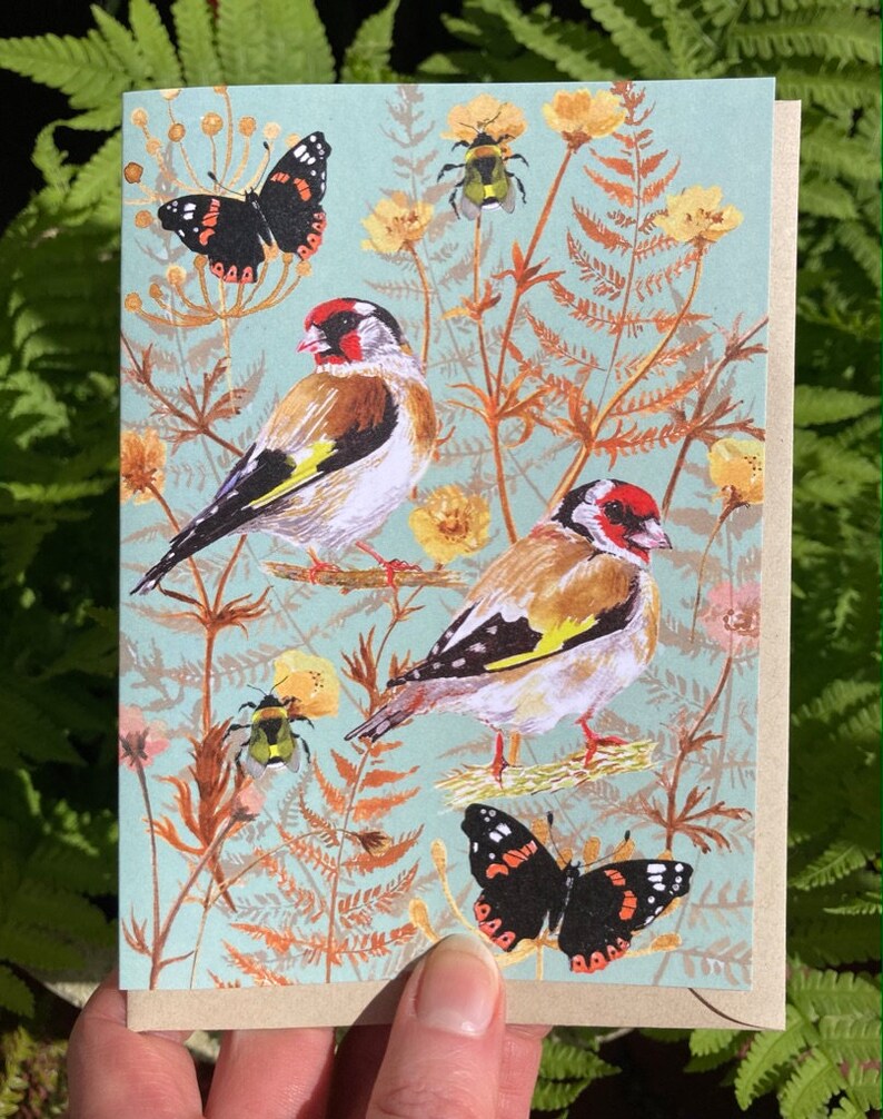 May include: A greeting card with a blue background featuring two goldfinches, two butterflies, and a bumblebee. The birds are perched on branches with green leaves and brown stems. The butterflies are flying in the air. The bumblebee is on a flower.