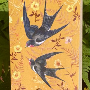 May include: A greeting card with a yellow background featuring two black and white swallows in flight. The background is filled with a floral pattern of yellow flowers and brown leaves.